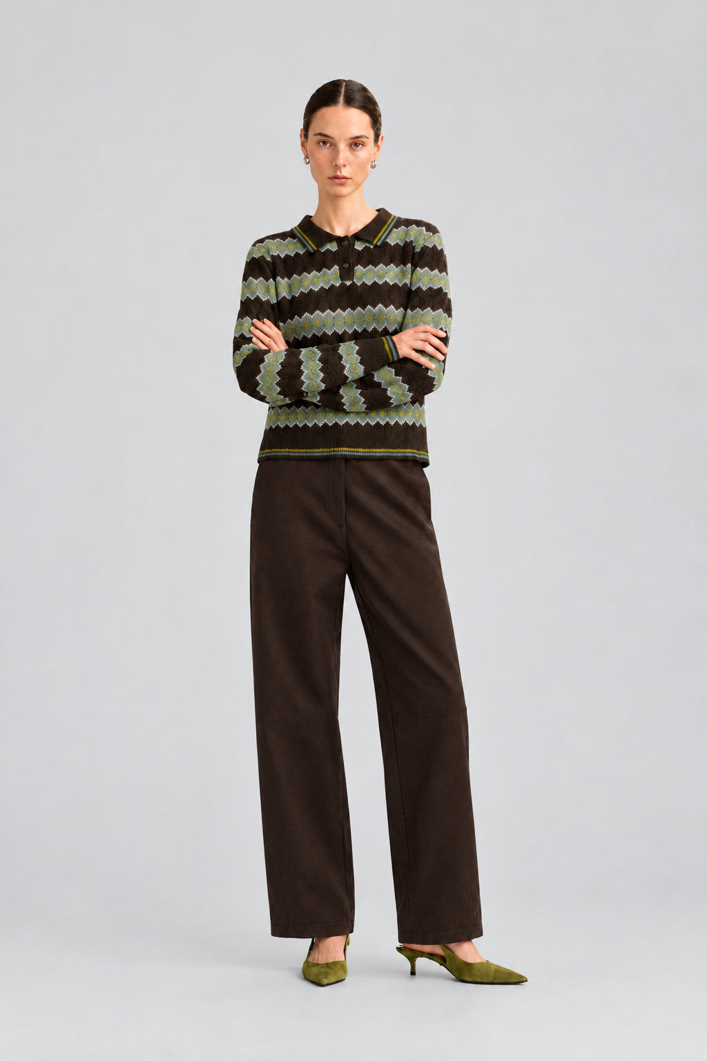 A model stands against a plain light background wearing a dark knit sweater with a geometric pattern in olive green, brown, and black, paired with dark brown wide-leg trousers and olive green pointed-toe heels.