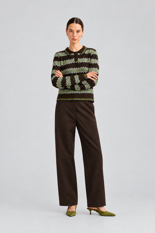A model stands against a plain light background wearing a dark knit sweater with a geometric pattern in olive green, brown, and black, paired with dark brown wide-leg trousers and olive green pointed-toe heels.